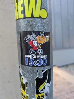 Street sticker The sticker is black and features a cartoon illustration of a bee dressed in a football kit (red shorts, white shirt with red trim, black and white football boots). The bee has orange skin, a large smile, and is holding a foamy pint of beer in its right hand. A soccer ball is depicted near the bee's feet. Below the illustration, the text 'ENDLICH WIEDER' is written in white, followed by '15:30' in large blue numerals with white outlines. The sticker appears to be applied to a metal pole and shows some signs of wear around the edges.