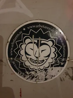 Street sticker This is a circular sticker with a black and white illustration of a stylized lion's face. The lion has a fierce expression with sharp teeth. The background of the sticker is black, with white text circling the illustration that reads "www.arbetaren.se". The sticker shows signs of wear and tear.