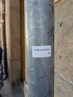 The sticker is a small, rectangular piece of white paper or vinyl, adhered to a textured, grey metal pole. The text is printed in black, sans-serif font, centered on the sticker. The sticker itself is slightly off-white due to lighting or age, and shows minor imperfections like slight creasing or uneven adhesion, particularly visible along its edges. The pole is scratched and worn, providing a stark contrast to the clean white of the sticker.