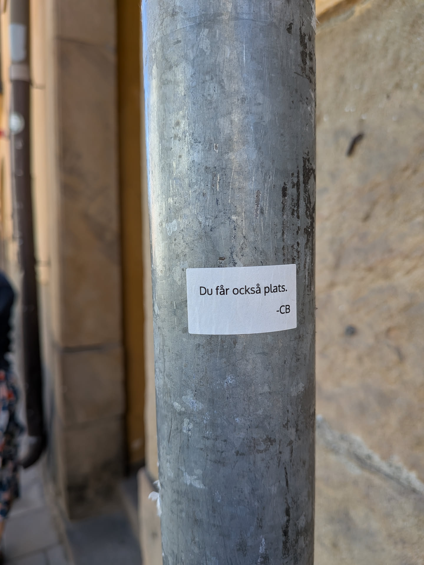 The sticker is a small, rectangular piece of white paper or vinyl, adhered to a textured, grey metal pole. The text is printed in black, sans-serif font, centered on the sticker. The sticker itself is slightly off-white due to lighting or age, and shows minor imperfections like slight creasing or uneven adhesion, particularly visible along its edges. The pole is scratched and worn, providing a stark contrast to the clean white of the sticker.