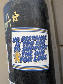 Street sticker The sticker is rectangular with a white background, featuring bold, blue text with a subtle shadow effect. The main message reads 'NO DISTANCE IS TOO FAR TO SUPPORT THE ONE YOU LOVE'. Below this, in a smaller, elegant script, it says 'Till minne av Åke'. To the left of the text, there's an illustration of a yellow lion, standing on its hind legs, wearing a crown, holding a sword, and carrying a shield adorned with three crowns. The top and bottom edges of the sticker are bordered by a pattern of thin blue and yellow horizontal stripes, with a wider blue stripe at the very edge. The sticker is adhered to a dark, weathered pole.