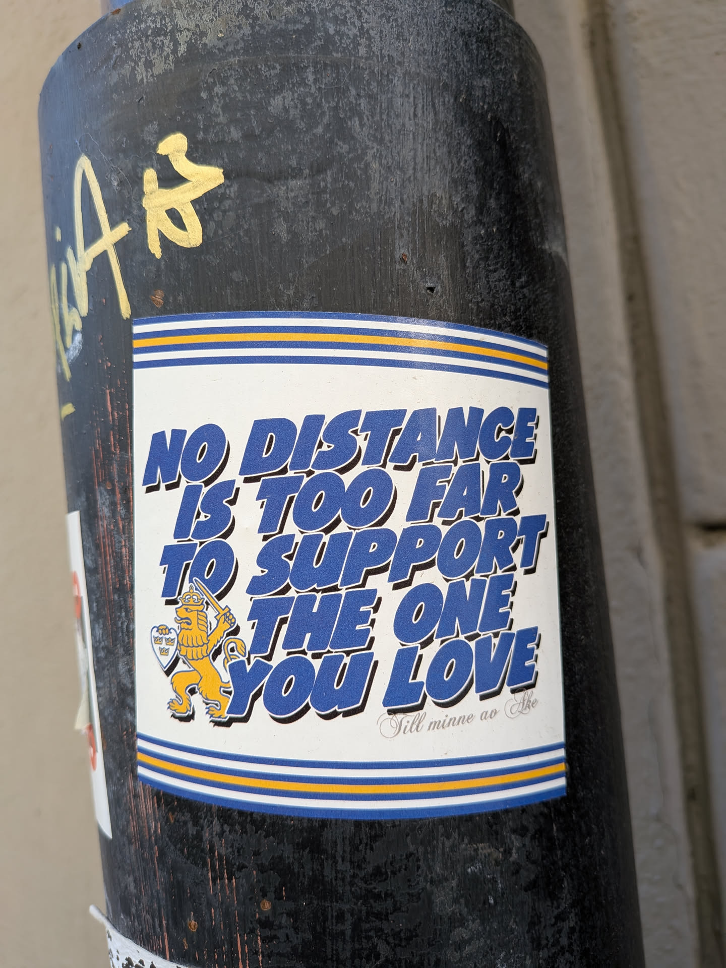 The sticker is rectangular with a white background, featuring bold, blue text with a subtle shadow effect. The main message reads 'NO DISTANCE IS TOO FAR TO SUPPORT THE ONE YOU LOVE'. Below this, in a smaller, elegant script, it says 'Till minne av Åke'. To the left of the text, there's an illustration of a yellow lion, standing on its hind legs, wearing a crown, holding a sword, and carrying a shield adorned with three crowns. The top and bottom edges of the sticker are bordered by a pattern of thin blue and yellow horizontal stripes, with a wider blue stripe at the very edge. The sticker is adhered to a dark, weathered pole.