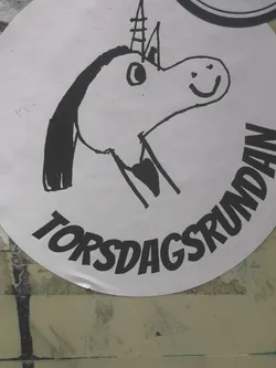 Street sticker A circular white sticker featuring a simple, hand-drawn cartoon unicorn head in black outline. The unicorn has a happy expression with a smiling mouth and a simple eye. It sports a pointed horn with horizontal line details and a flowing mane on its left side. Below the unicorn, the text "TORSDAGSRUNDAN" is written in bold, black, slightly slanted capital letters, following the curve of the sticker's bottom edge. The overall style is whimsical and childlike.
