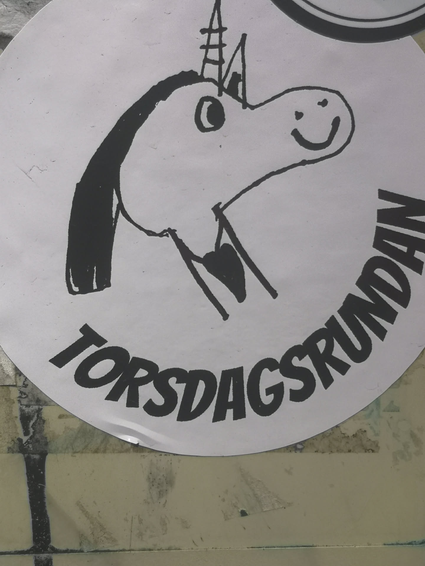 A circular white sticker featuring a simple, hand-drawn cartoon unicorn head in black outline. The unicorn has a happy expression with a smiling mouth and a simple eye. It sports a pointed horn with horizontal line details and a flowing mane on its left side. Below the unicorn, the text "TORSDAGSRUNDAN" is written in bold, black, slightly slanted capital letters, following the curve of the sticker's bottom edge. The overall style is whimsical and childlike.