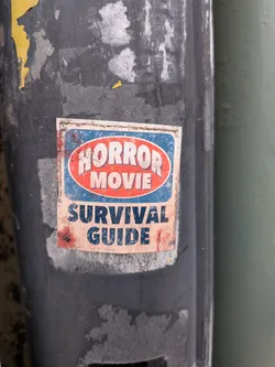 A rectangular sticker with a distressed, aged appearance, adhered to a dark, weathered pole. The top portion features an oval design with a red background and a white border, containing the words "HORROR MOVIE" in bold, white, sans-serif capital letters. This oval is set against a blue background with a white border. Below this, on a light beige/off-white background, are the words "SURVIVAL GUIDE" in bold, dark blue, sans-serif capital letters. The entire sticker shows significant wear, including dirt, scuffs, and reddish-brown smudges resembling bloodstains, particularly on the left side and bottom right. Its edges are slightly peeled and dirty, indicating exposure to the elements.