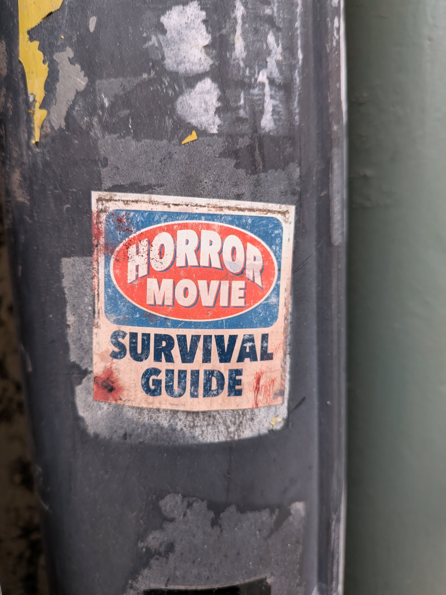 A rectangular sticker with a distressed, aged appearance, adhered to a dark, weathered pole. The top portion features an oval design with a red background and a white border, containing the words "HORROR MOVIE" in bold, white, sans-serif capital letters. This oval is set against a blue background with a white border. Below this, on a light beige/off-white background, are the words "SURVIVAL GUIDE" in bold, dark blue, sans-serif capital letters. The entire sticker shows significant wear, including dirt, scuffs, and reddish-brown smudges resembling bloodstains, particularly on the left side and bottom right. Its edges are slightly peeled and dirty, indicating exposure to the elements.