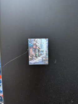 Street sticker The sticker is a small, square image depicting a detailed, illustrative scene of a narrow street or alleyway, reminiscent of an Asian city. The perspective draws the viewer down the street, which is flanked by multi-story buildings on both sides. These buildings feature various shopfronts with awnings, some in shades of blue and brown. Numerous red lanterns are strung across the street and hang from the building facades, adding pops of color. Potted plants and wooden benches are visible outside some establishments, contributing to a lively, lived-in atmosphere. The art style appears to be a detailed digital painting or illustration, characterized by a soft, somewhat muted color palette dominated by blues, browns, and reds. The ground is paved, and the overall impression is one of depth and a cozy, inviting urban environment.