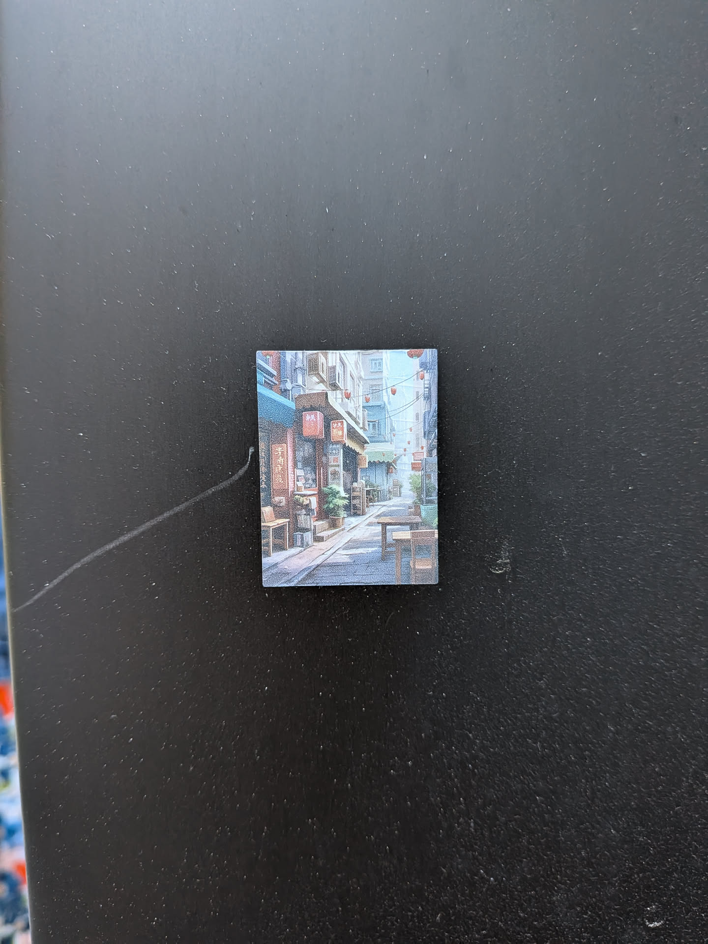 The sticker is a small, square image depicting a detailed, illustrative scene of a narrow street or alleyway, reminiscent of an Asian city. The perspective draws the viewer down the street, which is flanked by multi-story buildings on both sides. These buildings feature various shopfronts with awnings, some in shades of blue and brown. Numerous red lanterns are strung across the street and hang from the building facades, adding pops of color. Potted plants and wooden benches are visible outside some establishments, contributing to a lively, lived-in atmosphere. The art style appears to be a detailed digital painting or illustration, characterized by a soft, somewhat muted color palette dominated by blues, browns, and reds. The ground is paved, and the overall impression is one of depth and a cozy, inviting urban environment.
