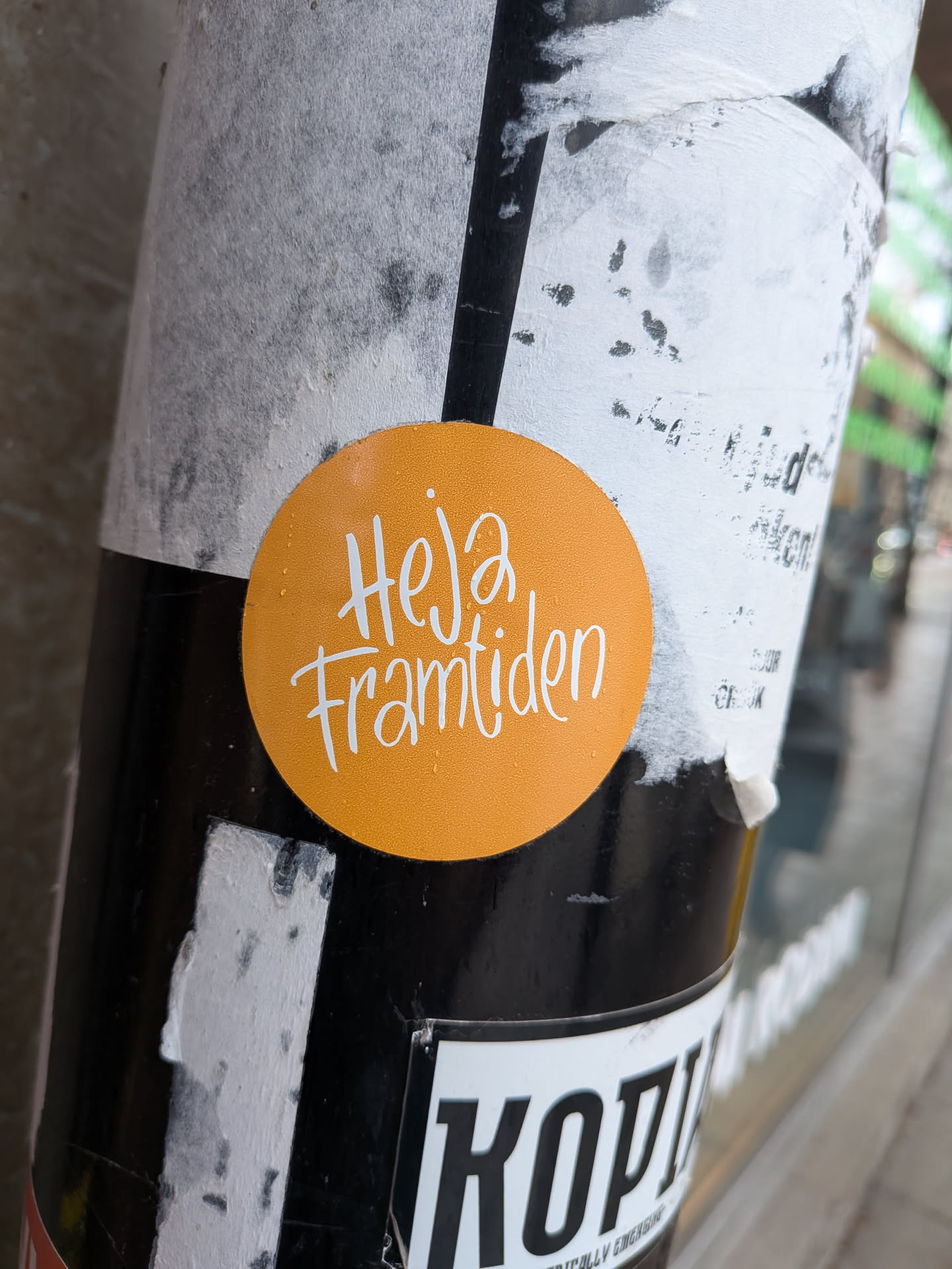 The sticker is perfectly circular with a bright, solid orange background. Centered on the sticker is the phrase 'Heja framtiden' written in a flowing, handwritten-style script font in white. The letters have a friendly, informal appearance. The surface of the sticker shows minor imperfections, possibly small water droplets or dust, and a slight sheen from ambient light. It is adhered to a textured, multi-layered pole covered with other torn paper and black sections.