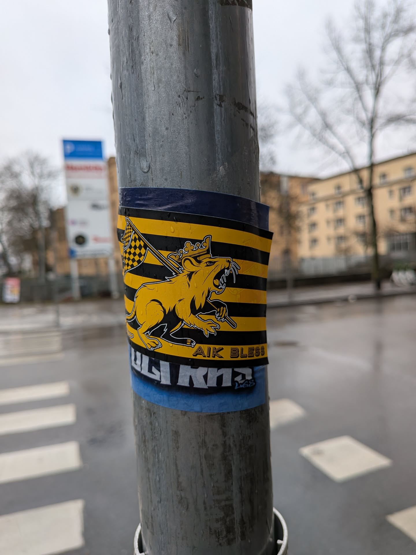 The sticker is rectangular, wrapped around a metal pole, and features a bold design of horizontal black and yellow stripes. Dominating the center is a stylized, yellow, lion-like creature with black outlines, depicted in a roaring or aggressive stance. It wears a crown on its head and appears to be holding a staff or scepter. Behind the creature, a black and yellow checkered flag is visible on a pole. Below the main graphic, the text "AIK BLESS" is clearly visible in white letters, and further down, partially obscured, the letters "ULTR" can be seen. The sticker shows signs of wear and is wet with water droplets.