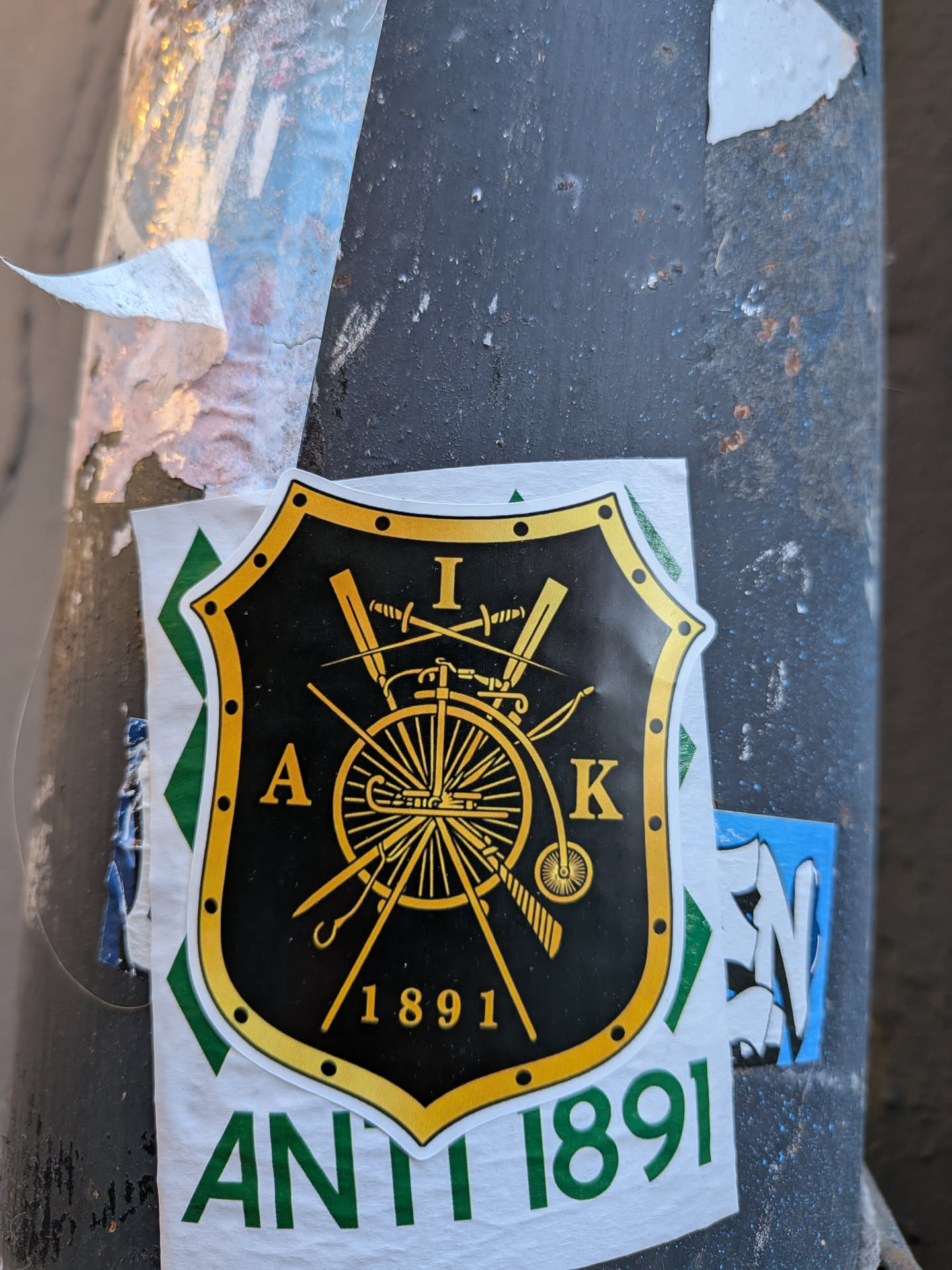The central sticker features a prominent black shield with a thick, ornate gold border, studded with small black circles. Inside the shield, various gold symbols are arranged: the letter 'I' at the top, 'A' on the left, and 'K' on the right. The main motif is a large spoked wheel, possibly from a penny-farthing bicycle or a wagon, crossed by two swords and other implements like a bow and an arrow or a pickaxe. Below these symbols, the year '1891' is displayed in gold. The entire shield design is placed on a white background, which extends below the shield to reveal the text 'ANTI 1891' in bold green capital letters. The sticker itself appears slightly worn and is affixed to a dark, textured pole, with other torn paper and stickers visible around it.