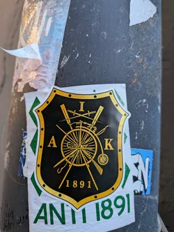 Street sticker The central sticker features a prominent black shield with a thick, ornate gold border, studded with small black circles. Inside the shield, various gold symbols are arranged: the letter 'I' at the top, 'A' on the left, and 'K' on the right. The main motif is a large spoked wheel, possibly from a penny-farthing bicycle or a wagon, crossed by two swords and other implements like a bow and an arrow or a pickaxe. Below these symbols, the year '1891' is displayed in gold. The entire shield design is placed on a white background, which extends below the shield to reveal the text 'ANTI 1891' in bold green capital letters. The sticker itself appears slightly worn and is affixed to a dark, textured pole, with other torn paper and stickers visible around it.