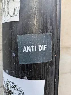 Street sticker A dark-green square sticker with the text "ANTI DIF" in white, uppercase letters. The sticker is affixed to a dark-colored pole, which also has other stickers on it.