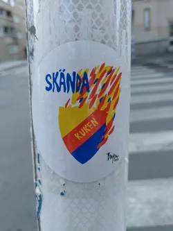 A round sticker with the text "SKÄNDA" in blue and yellow lettering. In the middle is a shield-shaped graphic with the text "KUKKEN" inside in red. The shield is surrounded by flames in red and orange. There's a small artist signature at the bottom.