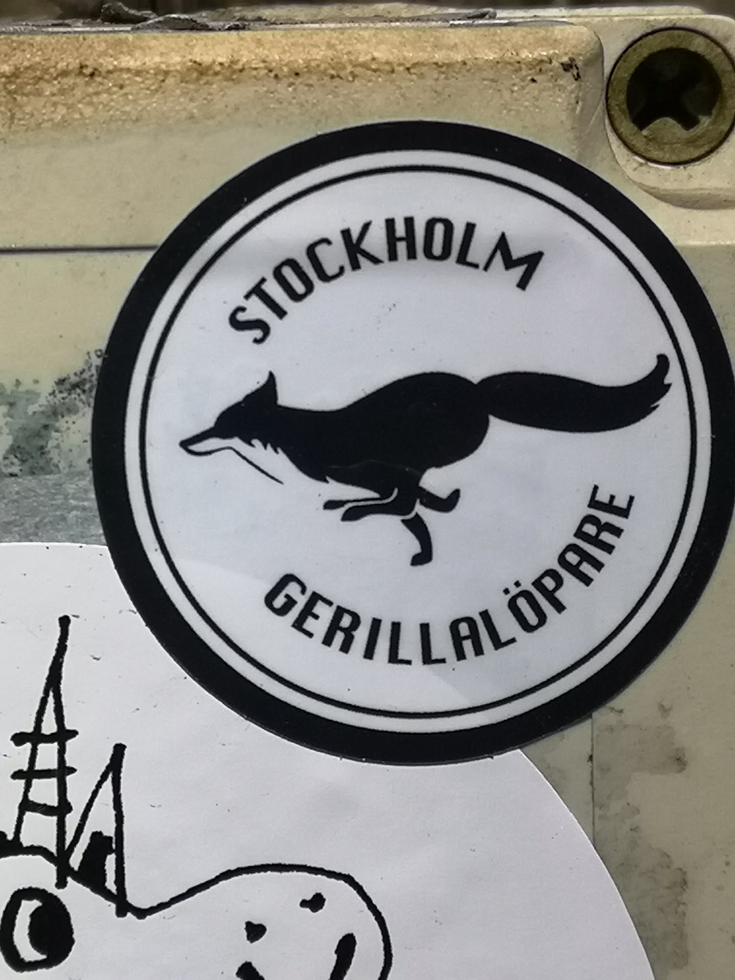 A circular sticker featuring a minimalist black and white design. It has a thick black outer ring enclosing a white background. Centered within the circle is a stylized black silhouette of a fox in a dynamic running or leaping pose, facing left with its tail extended. Curved along the top inner edge of the circle is the word 'STOCKHOLM' in black uppercase letters, and along the bottom inner edge is 'GERILLALÖPARE', also in black uppercase letters.