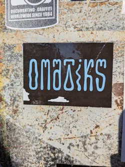 Street sticker A rectangular sticker featuring the word 'OMATIKS' in a distinctive light blue, bubble-letter style font. The letters are thick, rounded, and have a slightly glossy appearance. The background of the sticker is a dark, almost black or very dark brown, with some visible texture and reflections. There is minor wear and peeling at the bottom left edge, revealing white underneath. The sticker is adhered to a heavily rusted and textured metal surface.