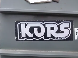 A black and white sticker with the word KORS in a stylized font.  Below the main text is a smaller line that says "RON'S". The sticker is rectangular and has a slightly worn appearance.