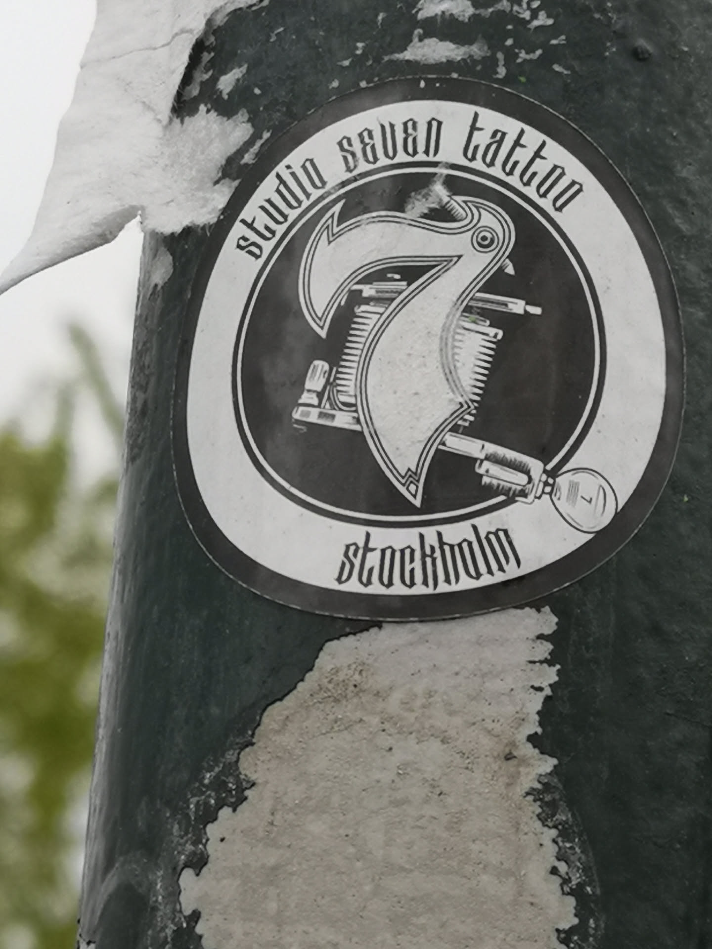 A circular, monochrome sticker featuring a stylized design for 'Studio Seven Tattoo'. The central element is a large, ornate number '7', which appears to be integrated with or overlaid on a classic coil tattoo machine. The text 'studio seven tattoo' is curved along the top edge of the circle, and 'stockholm' is curved along the bottom edge, both rendered in a distinctive gothic or old English script. The overall aesthetic is vintage and bold, presented in black and white tones. There's also a small '7' visible within a circular detail on the tattoo machine's grip.