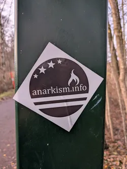 Street sticker The sticker is square-shaped and rotated 45 degrees, appearing as a diamond. It features a dark brown circular emblem with a white border. Inside the circle, at the top, are four white stars arranged in an arc. Below the stars, the text 'anarkism.info' is written in white, sans-serif font. Beneath the text are two thick white horizontal lines, with a curved white shape resembling a flame positioned above the left side of the text. The sticker has a glossy finish and is adhered to a dark green, textured surface.