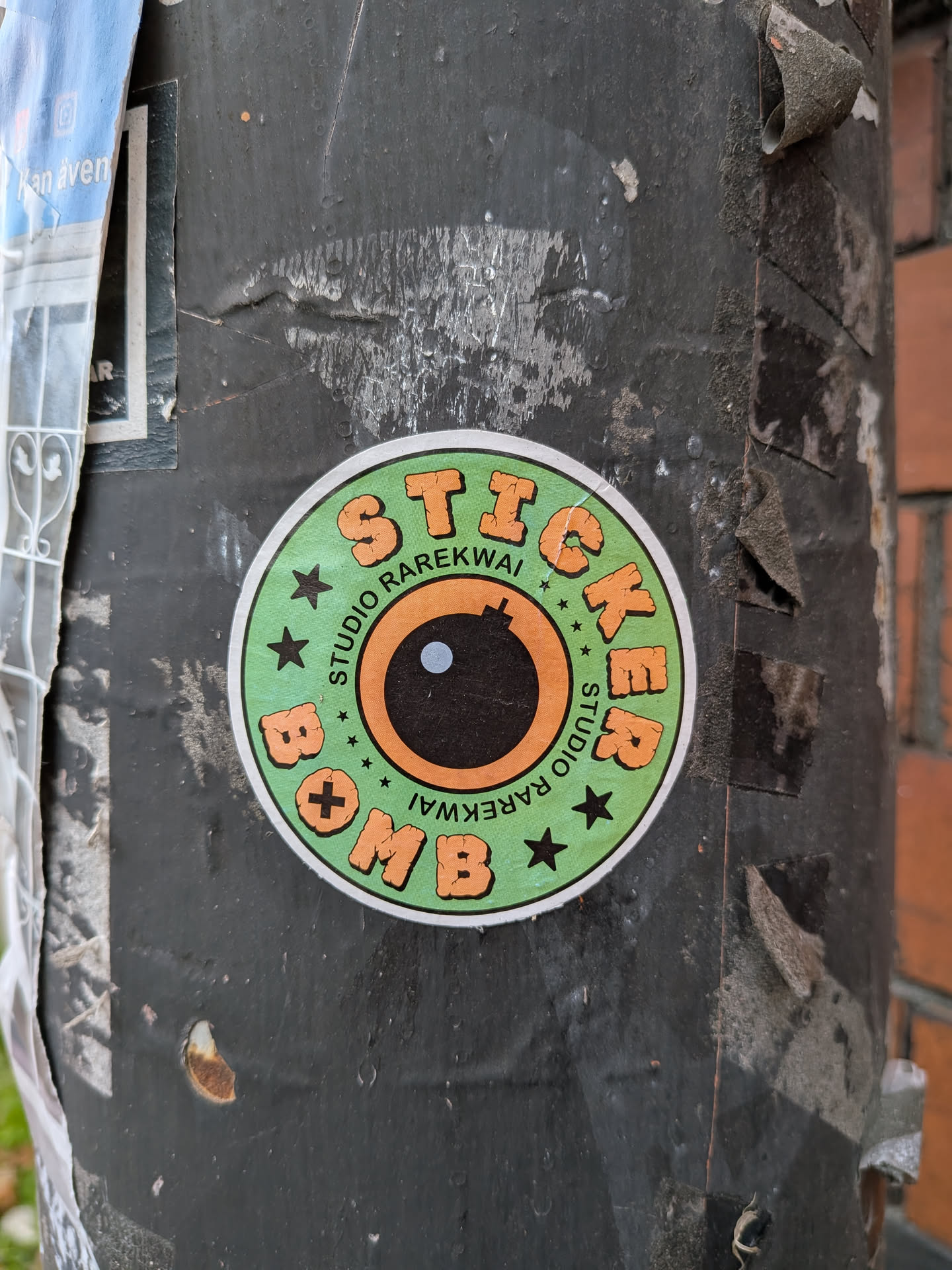 The sticker is circular with a white border. The background is a light green color with small black stars and plus signs scattered around. In the center is a stylized representation of a bomb's fuse and eye, with an orange ring around a black circle that has a small white dot in its center. The words 'STUDIO RAREKWAI' are written in a curve around the top half of the bomb's eye, and 'STICKER BOMB' is written in large, orange, blocky letters around the bottom half, with the letters 'BOMB' appearing upside down. The letters have a textured, slightly distressed appearance.