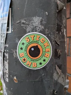 Street sticker The sticker is circular with a white border. The background is a light green color with small black stars and plus signs scattered around. In the center is a stylized representation of a bomb's fuse and eye, with an orange ring around a black circle that has a small white dot in its center. The words 'STUDIO RAREKWAI' are written in a curve around the top half of the bomb's eye, and 'STICKER BOMB' is written in large, orange, blocky letters around the bottom half, with the letters 'BOMB' appearing upside down. The letters have a textured, slightly distressed appearance.