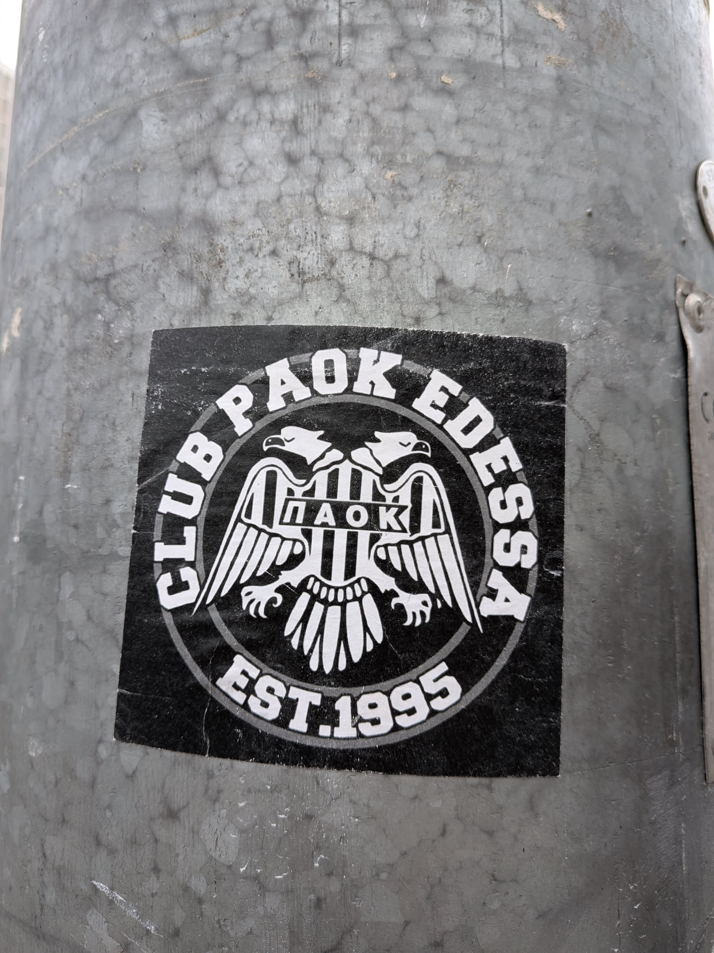 A circular, black and white sticker featuring a stylized double-headed eagle as its central motif. The eagle has outstretched wings and holds a shield on its chest, which displays vertical black and white stripes and the Greek letters 'ΠΑΟΚ' (PAOK). Encircling the eagle, in an arc at the top, is the text 'CLUB PAOK EDESSA' in white capital letters. Below the eagle, in a similar arc, is 'EST.1995'. The entire design is bordered by a white and grey double ring. The sticker is applied to a textured grey metal pole and shows minor signs of wear and creasing.