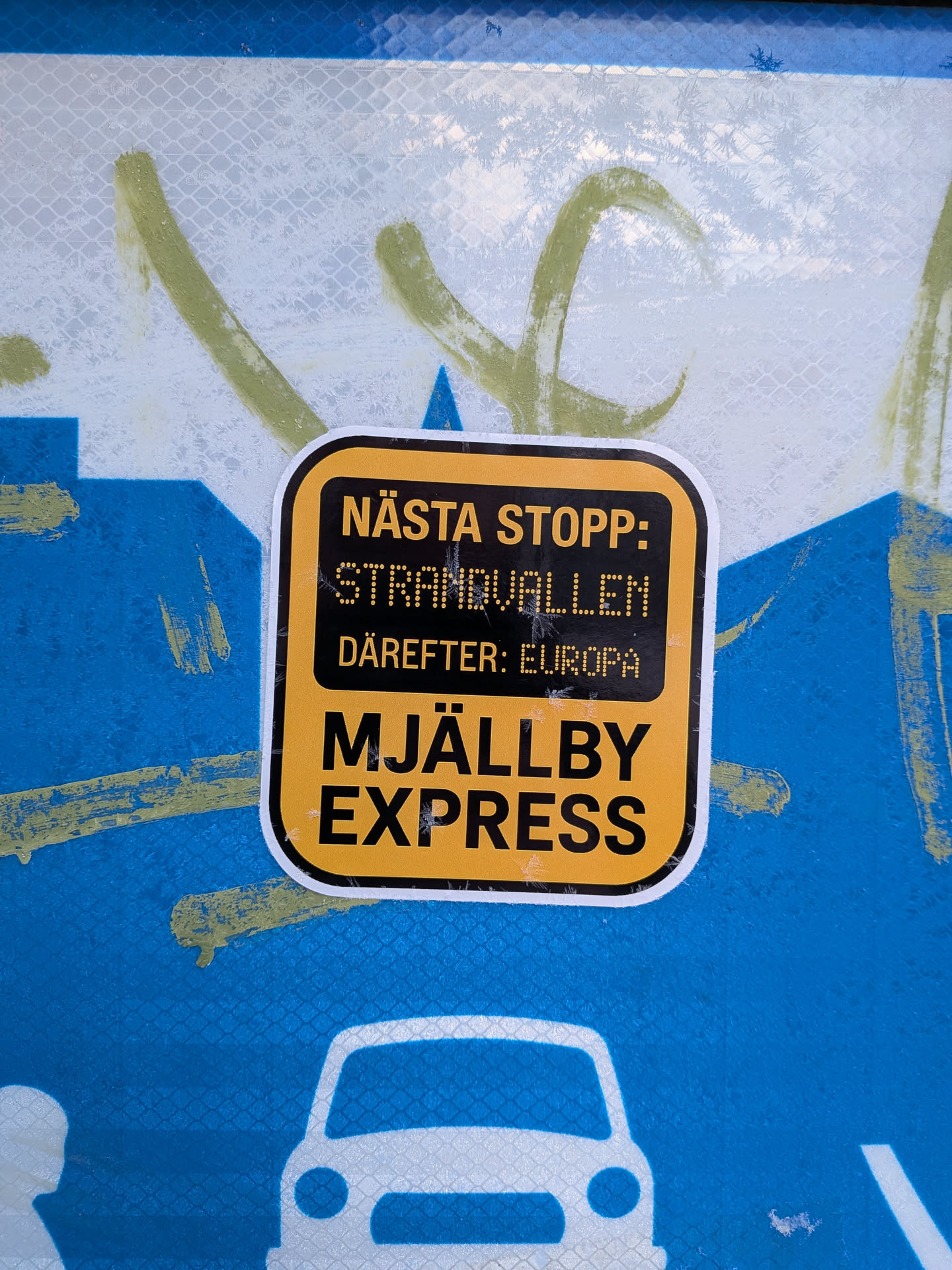 Street sticker A rectangular sticker with rounded corners, featuring a bold yellow and black color scheme. The top and bottom sections have black text on a yellow background, while the middle section has yellow text on a black background. The text 'STRANDVALLEN' and 'EUROPA' appear in a pixelated, dot-matrix style font, mimicking an old-school departure board. The sticker has a thin white border around its edge and is applied to a textured, reflective blue and white surface with green graffiti.