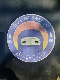 Street sticker Round sticker featuring a cute cartoon character wearing a purple hood. The sticker has a pastel color scheme and the text "Seal of Sweden" at the top and "Bättre Lycka" at the bottom. The text is in a stylized font and the character has a melancholic expression.
