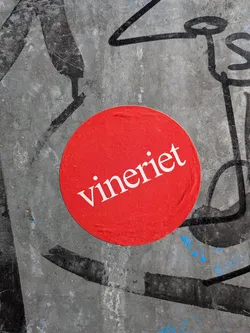 Street sticker The sticker is a perfectly circular, vibrant red disc, centrally placed on a grimy, dark grey surface covered with black graffiti lines and faint blue paint splatters. The sticker itself has a slightly textured, matte appearance with minor creases, indicating it's a physical object. In the center of the red circle, the word 'vineriet' is written in a clean, white serif font, slightly angled upwards from left to right. The contrast between the bright red and white text makes it highly legible against the dark, chaotic background.