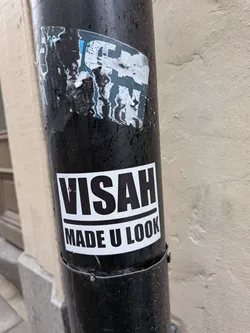 Street sticker A rectangular, monochrome sticker featuring bold black text on a white background. The top line reads 'VISAH' in large, sans-serif capital letters. Below it, separated by a thin black line, the second line reads 'MADE U LOOK' in similar but slightly smaller capital letters. The sticker is applied to a dark, possibly black, metal drainpipe, showing some minor wear and tear, particularly around the edges and a small scuff on the letter 'S' in 'VISAH'.