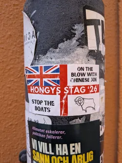 Street sticker A rectangular sticker prominently featuring the Union Jack flag on its left side. To the right of the flag, the text 'ON THE BLOW WITH CHINESE JOE' is displayed in black capital letters. Below the flag, the text 'HONGYS STAG '26' is written in large white capital letters against a red background, extending across the sticker. Further below, 'STOP THE BOATS' is printed in black capital letters on a white background. In the bottom right corner, there is a simple black outline drawing of a ram's head. The sticker shows signs of wear and tear, with some peeling and creasing, indicating it is a physical sticker affixed to a textured black pole.