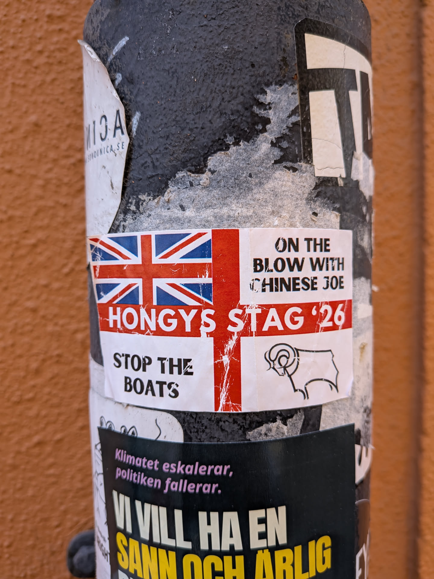 A rectangular sticker prominently featuring the Union Jack flag on its left side. To the right of the flag, the text 'ON THE BLOW WITH CHINESE JOE' is displayed in black capital letters. Below the flag, the text 'HONGYS STAG '26' is written in large white capital letters against a red background, extending across the sticker. Further below, 'STOP THE BOATS' is printed in black capital letters on a white background. In the bottom right corner, there is a simple black outline drawing of a ram's head. The sticker shows signs of wear and tear, with some peeling and creasing, indicating it is a physical sticker affixed to a textured black pole.