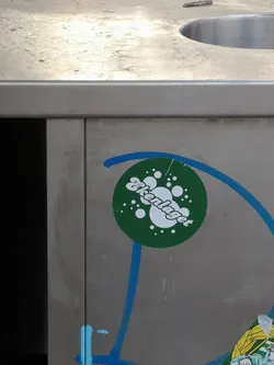 Street sticker The sticker is circular with a dark green background. It features white text in a playful, slightly cursive font that reads "Ekenlaget". Surrounding the text are numerous white circles of varying sizes, giving the impression of bubbles or effervescence. The sticker shows signs of wear, with a small tear at the top edge and some minor scuffs. It is affixed to a metallic, possibly stainless steel, surface, which also has blue spray paint marks partially overlapping the sticker.