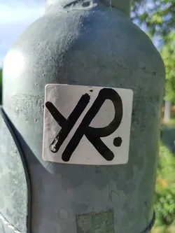 Close-up image of a square white sticker with black lettering. The sticker displays the letters "YR." written in a stylized, possibly graffiti-inspired font.  The sticker is affixed to a weathered gray metal pole outdoors.