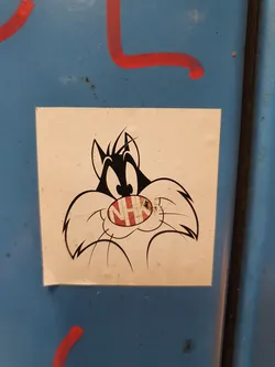 A sticker featuring Sylvester Jr. from Looney Tunes. The sticker is affixed to a blue surface with some red graffiti visible in the background. The sticker shows Sylvester's face with his signature mustache and a small red logo on his nose.