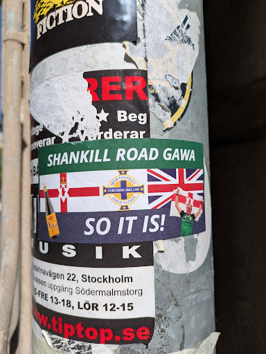 This sticker displays the text "SHANKILL ROAD GAWA" along with flags, a logo of the Northern Ireland association and a football supporter with the Union Jack and a bottle. Below it is the text "SO IT IS!". This sticker is placed on a metal pole on top of another sticker.