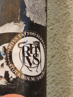 Street sticker A round sticker with a black and white illustration of a wolf-like creature encircling the letters RITVS. The text "Ritvs Tattoo Supplies Stockholm/Sweden" is written around the circular design. The sticker appears to be affixed to a dark-colored pole.