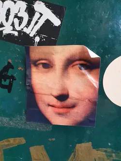 A sticker featuring a close-up of the Mona Lisa's face. The sticker has a slightly worn look, suggesting it may have been placed outdoors for some time. The background is a dark teal surface with other graffiti and stickers visible.