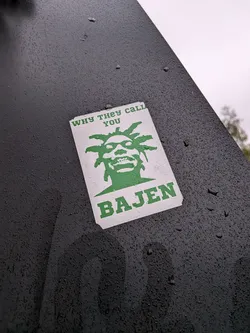 Street sticker A rectangular sticker featuring a stylized, almost cartoonish portrait of a person with dreadlocks and a wide, toothy grin, rendered in solid green against a white background. Above the portrait, the text 'WHY THEY CALL YOU' is written in green capital letters. Below the portrait, the name 'BAJEN' is prominently displayed in larger, bold green capital letters. The sticker is affixed to a dark, textured surface, possibly metal, which is covered in numerous small water droplets, as is the surface of the sticker itself, giving it a weathered, outdoor appearance.