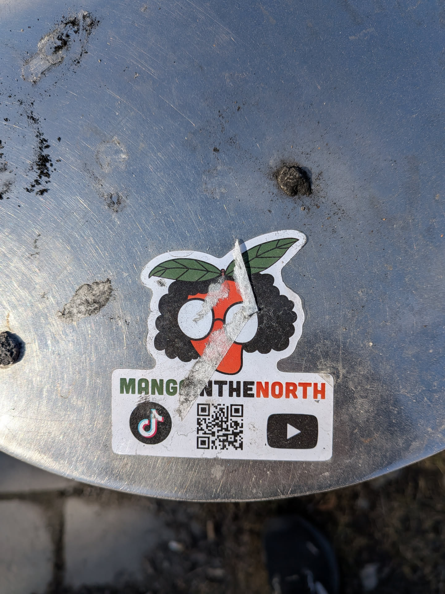 A vibrant sticker featuring a stylized, anthropomorphic mango character. The mango has an orange body, topped with two green leaves, and sports a black, curly afro-like hairstyle. It wears white-rimmed circular glasses. A diagonal tear runs across the character's face. Below the character, a white banner displays the text 'MANGONTHENORTH'. Further down, on the same white background, are a TikTok logo, a QR code, and a YouTube play button logo. The sticker is affixed to a scratched metallic surface and shows signs of wear and dirt.