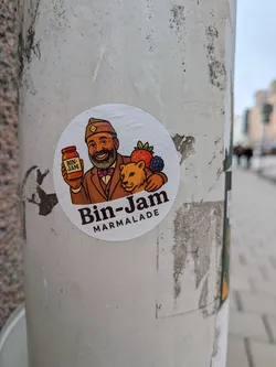 Street sticker The sticker is circular with a white background, featuring a colorful cartoon illustration. It depicts a smiling, bearded man wearing a brown uniform-style hat with a yellow square emblem, a brown suit jacket, and a purple bow tie. He holds up a small, orange-labeled jar that reads 'BIN-JAM'. To his right, a friendly-looking, light brown lion cub is shown, with a red strawberry and a dark blue blackberry resting on its back. Below the illustration, the words 'Bin-Jam' are prominently displayed in a large, bold, dark brown font, with 'MARMALADE' written beneath it in a smaller, dark brown, sans-serif font. The overall aesthetic is warm, inviting, and reminiscent of classic food branding.