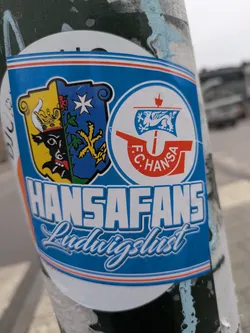 Street sticker A sticker displaying the coat of arms and logo of Hansa Fans Ludwigslust, a football club's fan group. The sticker features a blue and white design with the team's crest on the left and the club's logo on the right. The text 'HANSAFANS Ludwigslust' is prominently displayed at the bottom.