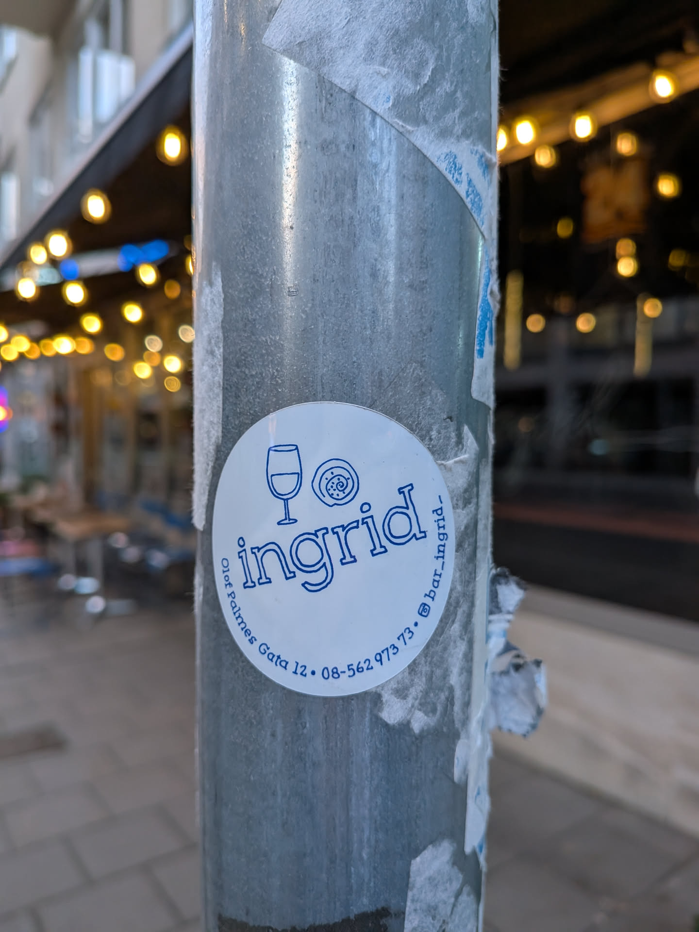 Street sticker A circular sticker with a white background and blue lettering and graphics. At the top left, there is a simple illustration of a wine glass. To its right is a stylized drawing of a pastry or bun. The word 'ingrid' is prominently displayed in the center in a bold, sans-serif font. Along the bottom edge, in smaller text, are the words 'Olof Palmes Gata 12' and a phone number '08-562 973 73'. To the right of the phone number, there is an Instagram logo followed by '@bar_ingrid_'.