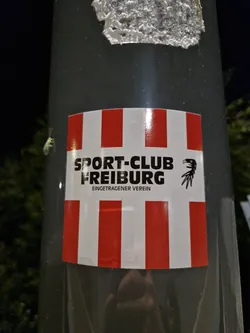 Street sticker A sticker featuring the logo of Sport-Club Freiburg, a German football club. The sticker has a red and white striped design and includes the text "Sport-Club Freiburg" and "eingetragener Verein" (registered association).