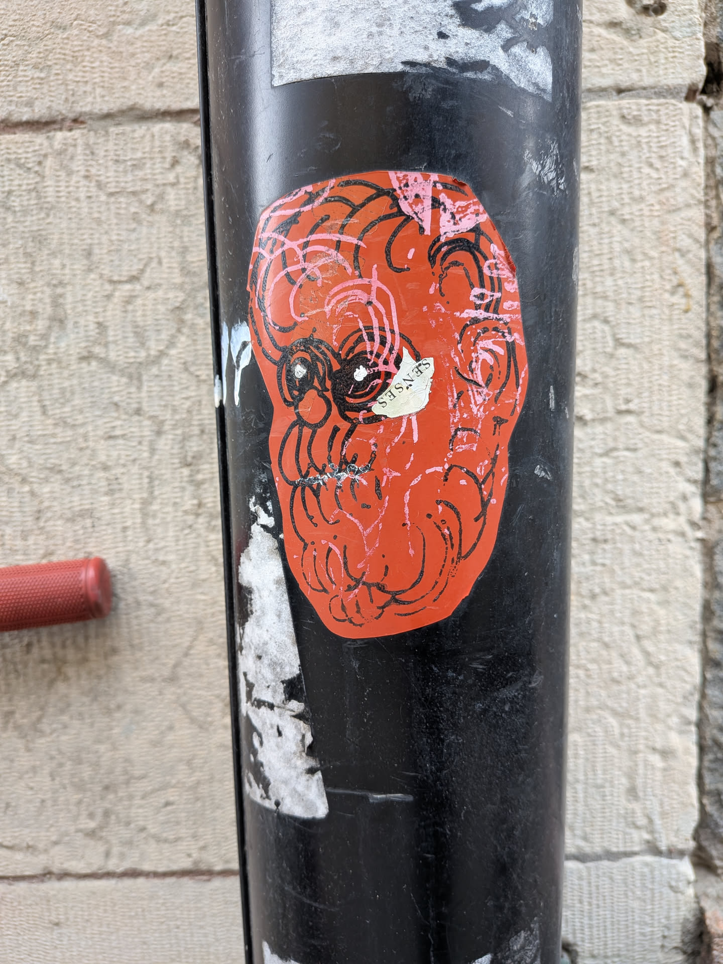 The sticker is roughly oval-shaped, resembling a stylized skull or face, set against a bright, somewhat faded orange background. It is affixed to a black metal pole, showing signs of wear and tear, including some peeling around the edges and minor scuffs on its surface. The design features intricate, swirling black lines that form the contours of the face, with prominent black circular eyes that have small white dots. Overlaid on these black lines are thinner, lighter pink lines, adding a layer of depth and a contrasting color. A small, torn white piece of paper or sticker is partially adhered over the left eye area, with the word 'SENSES' written vertically in black text. The overall aesthetic is one of raw, energetic urban art with a slightly distressed, hand-drawn feel.