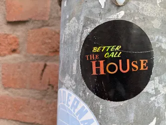 Street sticker A black round sticker on a grey metallic pole shows the text 'Better the call House' with the word House in orange.