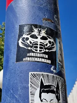 A black and white sticker of a muscular bull. The sticker is on a blue pole. The text on the sticker says #freebiffen #freeeharmano.