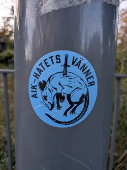 Street sticker The sticker is circular with a bright blue background. In the center, a black silhouette of a rat is depicted. The rat is curled up, with its head down and its tail forming a loop. A black sword is plunged vertically through the rat's back, with the hilt visible above the rat's body. The rat has its eyes crossed out with 'X' marks, and its fur is rendered with sharp, angular lines. The text 'AIK-HATETS VÄNNER' is written in black, uppercase letters in an arc around the top of the sticker.