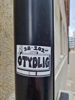 Street sticker This sticker is placed on a black pipe and features the text 'OTYDLIG' in large, bold, black letters on a white background. Above the text, there's a combination of numbers and a graphic resembling a pixelated face or logo. The sticker has a border reminiscent of a classic license plate design.