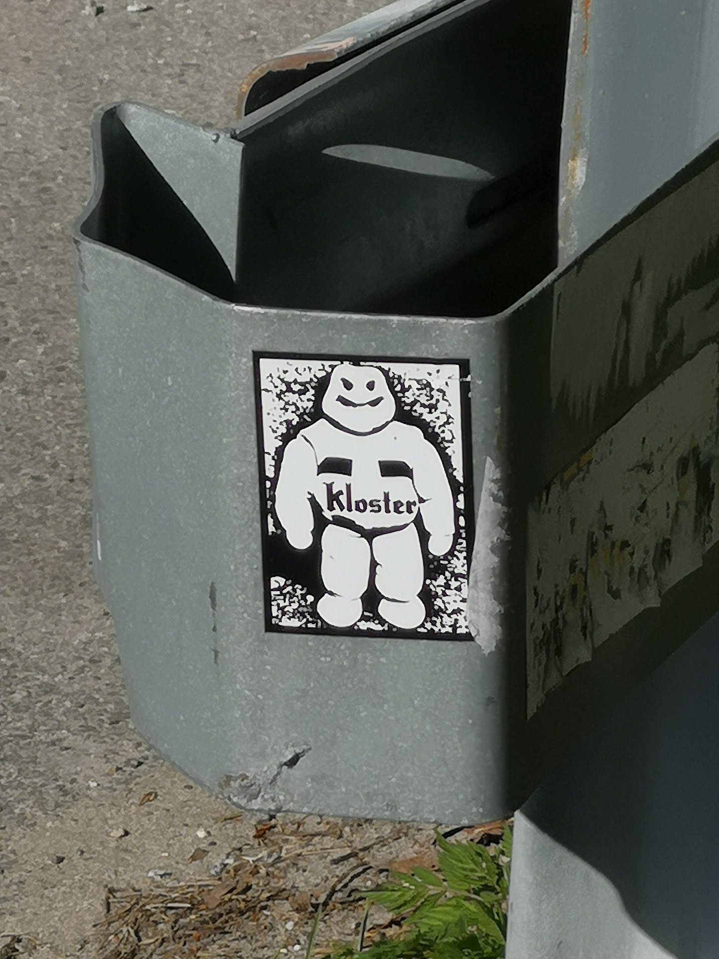 A rectangular, vertically oriented sticker featuring a stylized, portly white character with a simple, friendly smile and two dot eyes. The character has short, rounded limbs. Across its chest, a black band displays the word "Kloster" in a white, somewhat gothic or stylized font. The background behind the character is black with a white speckled or distressed texture, creating a high-contrast, gritty effect. The entire sticker is rendered in black and white.